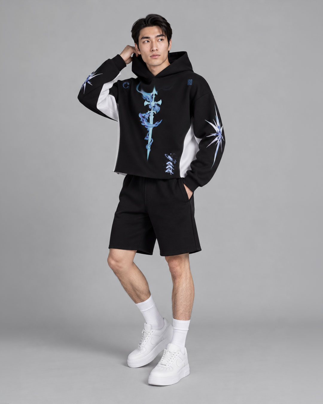 Model wearing a black hoodie with a blue mystical sword graphic on the chest and a starburst print on the sleeve, white side panels, paired with black shorts and white chunky sneakers — streetwear brand hoodie collection.