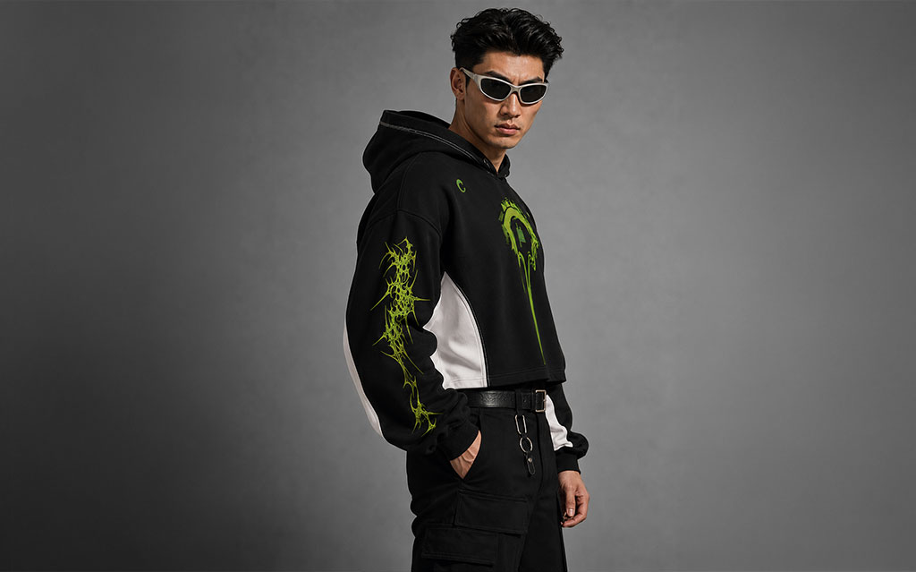 Back view of model wearing a black cropped hoodie with a neon green dragon graphic and skull design on the hood, matching thorn-vine sleeve prints, white side panels, paired with black cargo pants and a black belt — streetwear brand hoodie collection.