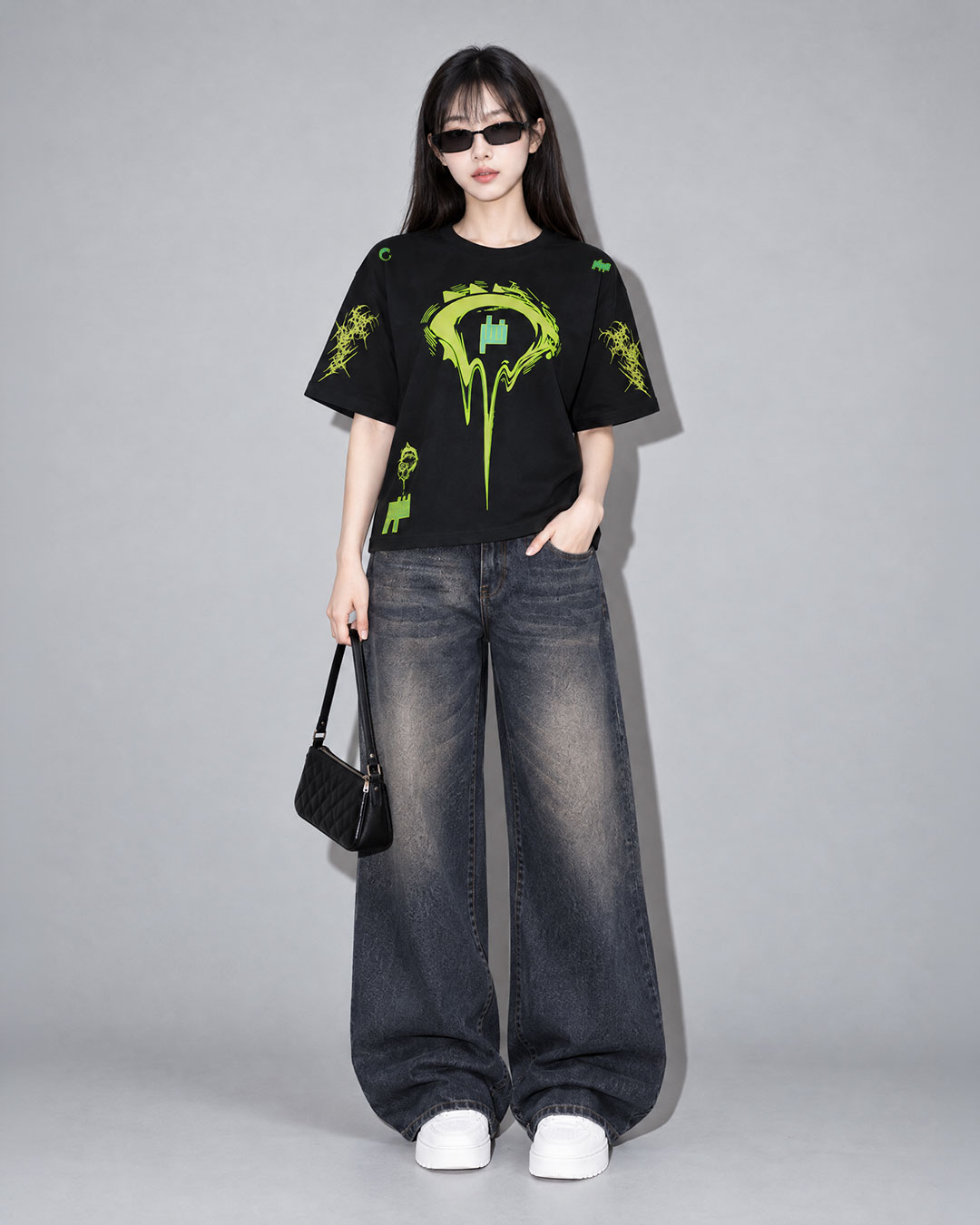 Female model in sunglasses wearing a black t-shirt with a neon green dripping eye graphic and scattered logo details, paired with dark wash wide-leg jeans, a black quilted shoulder bag, and white sneakers, neutral gray studio background.