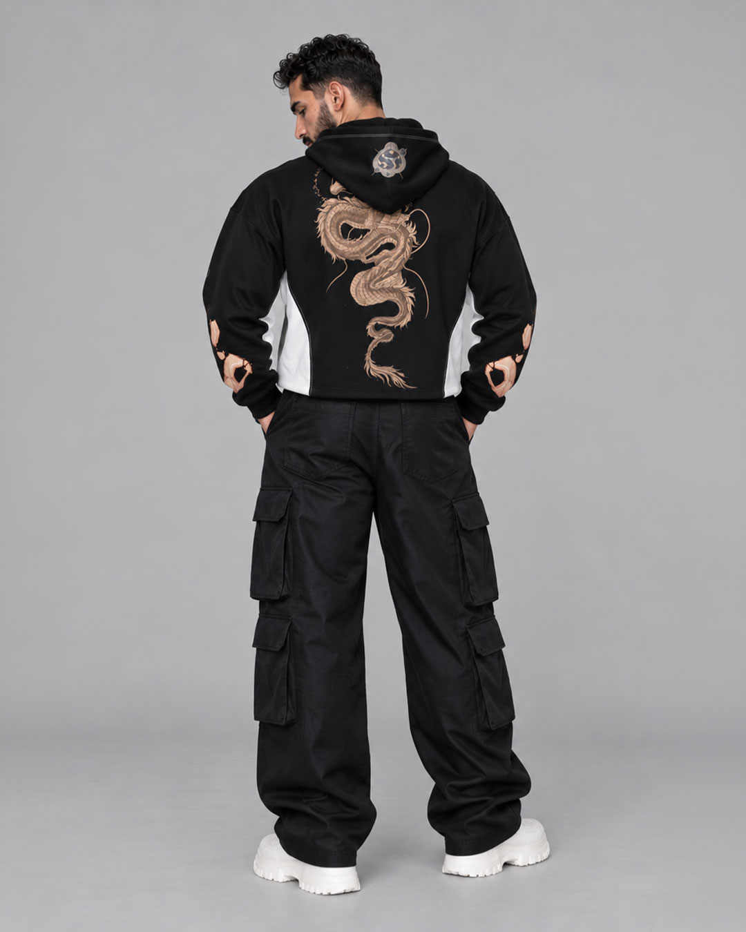 Back view of model wearing a black box-fit cropped hoodie with a large gold serpentine dragon print running down the spine and a yin-yang patch on the hood, paired with black wide-leg cargo pants and white chunky sneakers — streetwear brand hoodie collection.