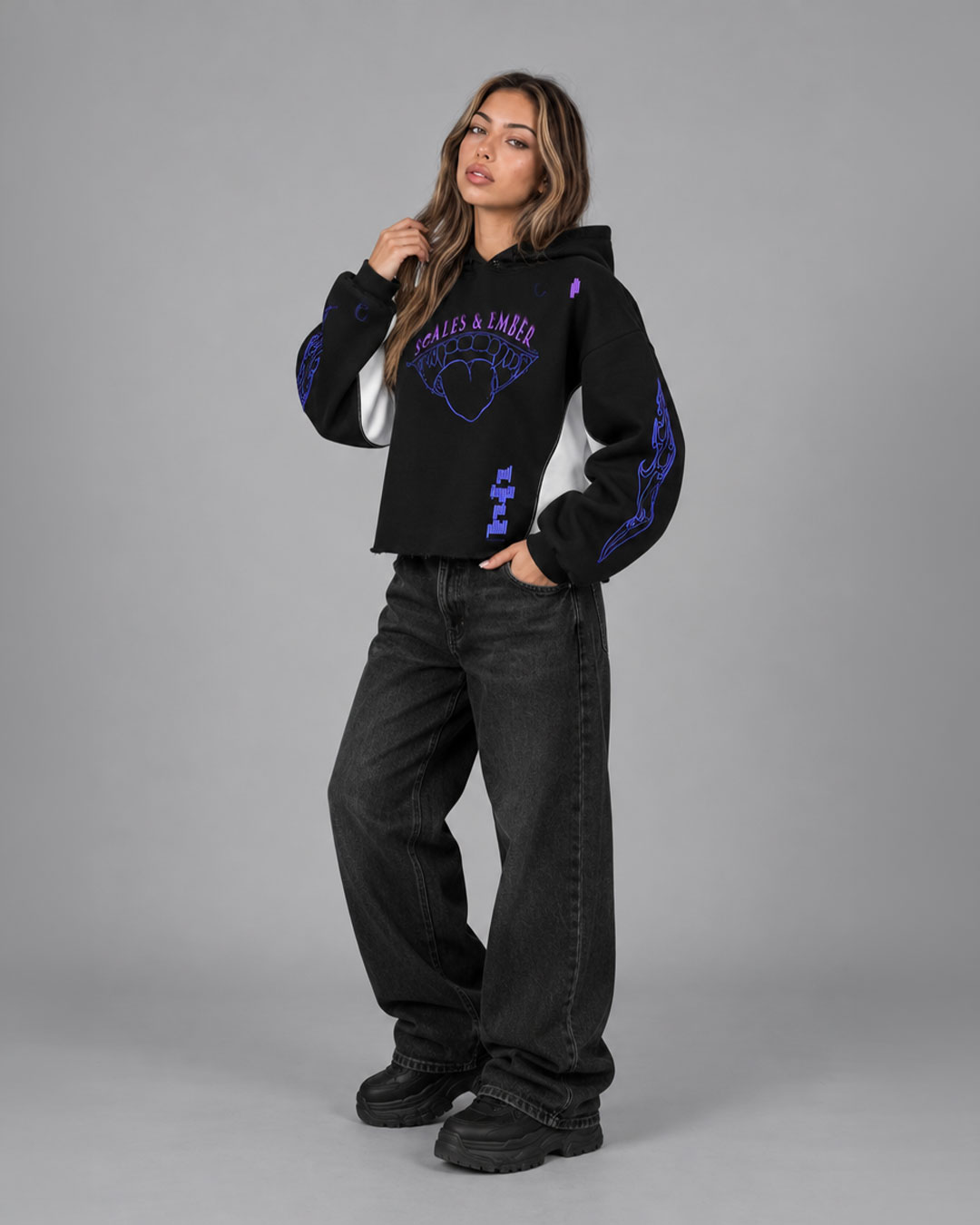 Model wearing a black cropped hoodie with a purple and blue dragon jaw graphic and "Scales & Ember" branding on the chest, flame sleeve print, paired with black wide-leg jeans and black chunky boots — streetwear brand hoodie collection.