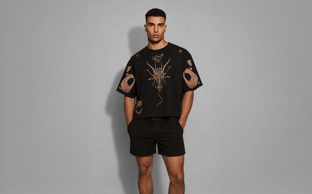 Model wearing a black oversized t-shirt with a gold dragon graphic and Arabic calligraphy print on the chest, hand tattoo sleeve prints, paired with black shorts — streetwear brand t-shirt collection.