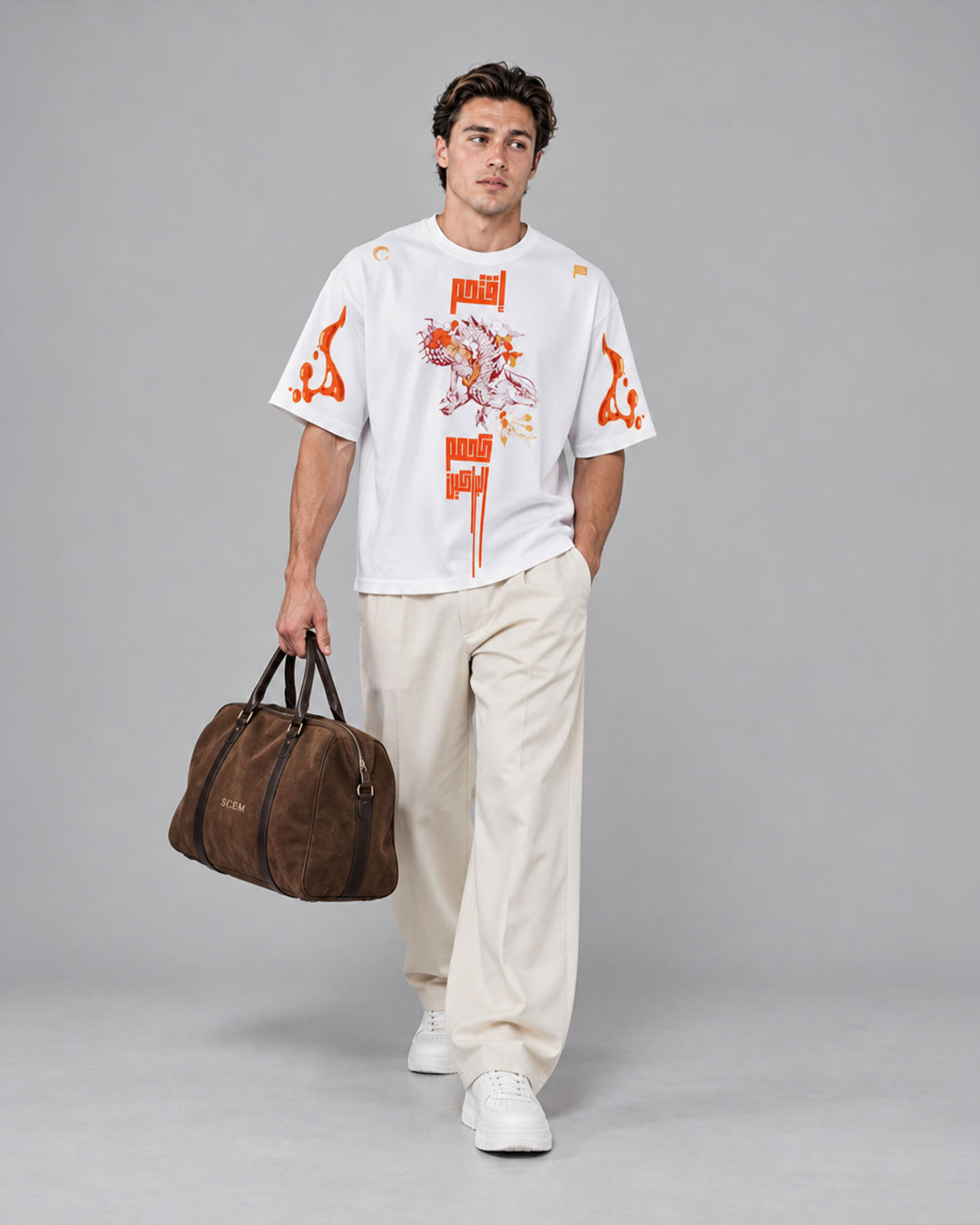 Model wearing a white t-shirt with an orange dragon graphic and Arabic calligraphy print, flame accents on the sleeves, paired with cream wide-leg trousers, a brown suede duffel bag, and white sneakers — streetwear brand t-shirt collection.