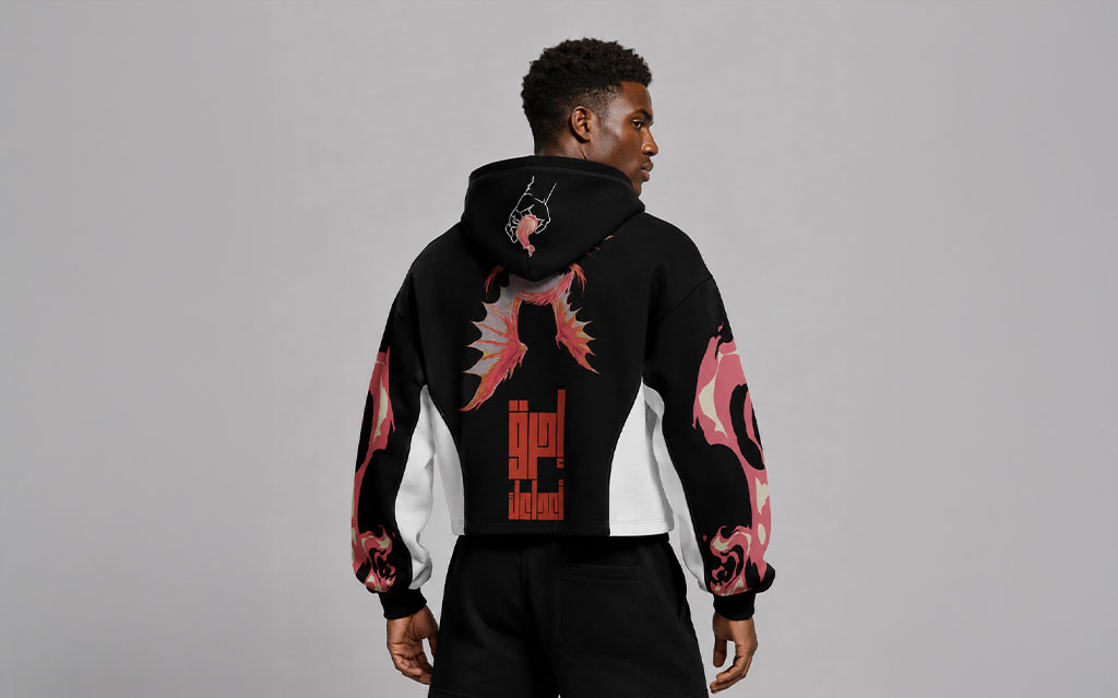 Back view of model wearing a black cropped hoodie with a pink and orange fire-wing graphic and red Arabic calligraphy print on the back, flame camo sleeve prints, white side panels, paired with black shorts — streetwear brand hoodie collection.