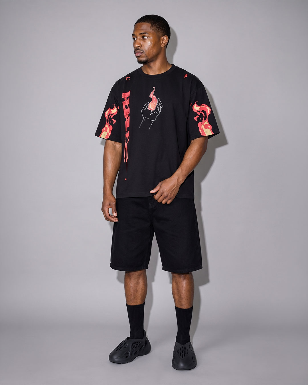 Male model wearing a black t-shirt with a hand-holding-flame graphic and red script text on the chest, matching flame sleeve prints, paired with black shorts, black socks, and black slide sneakers, neutral gray studio background.