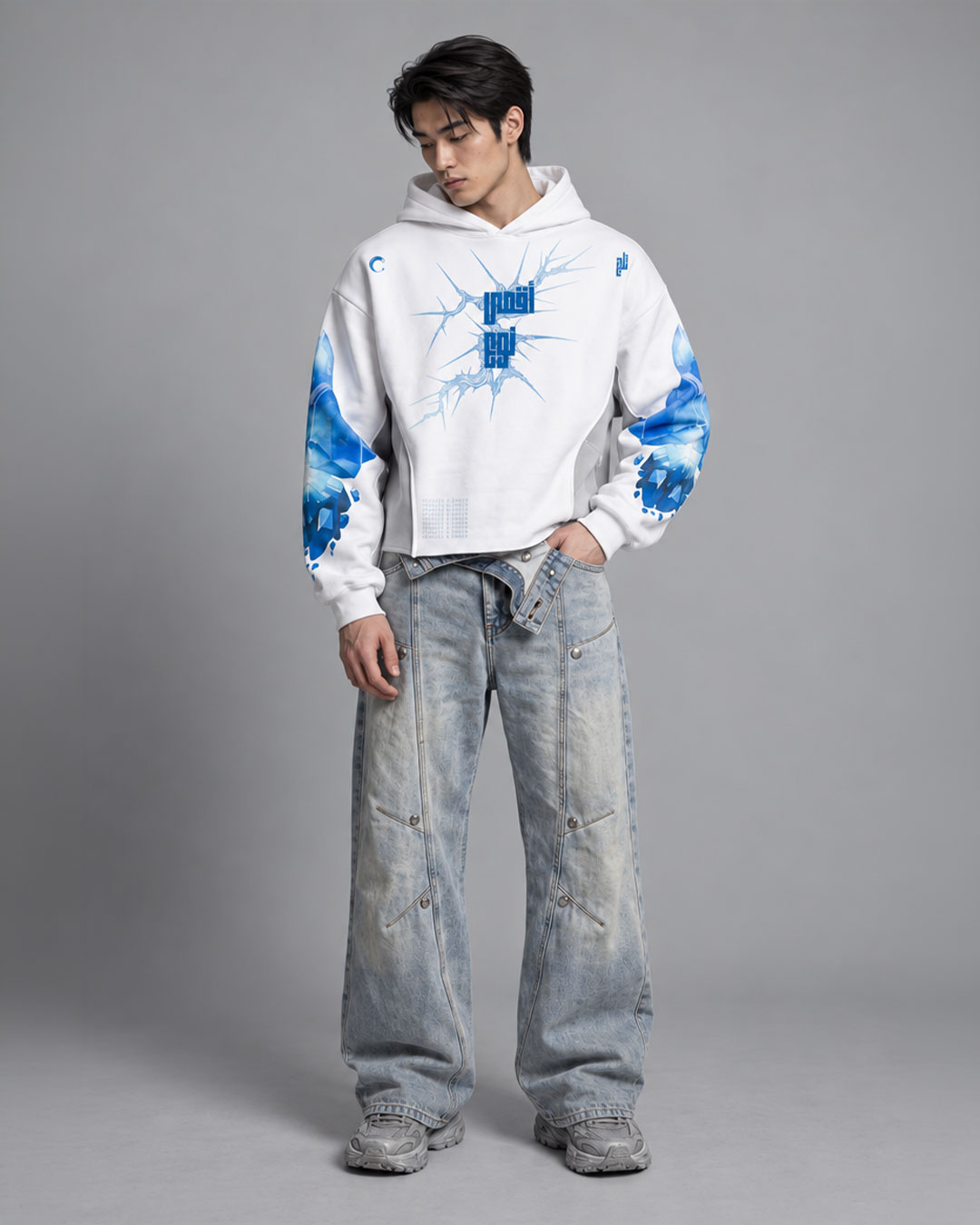 Front view of male model wearing a white cropped hoodie with a mechanical winged cross graphic print between the shoulder blades and blue crystal ice designs on both sleeves, paired with light-wash baggy jeans and gray sneakers, neutral studio background.