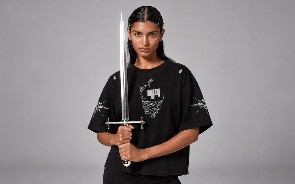 Female model holding a silver sword upright in front of her face, wearing a black cropped t-shirt with white graphic eye and tribal spike designs and a stylized script logo across the chest, paired with black bottoms, neutral gray studio background.