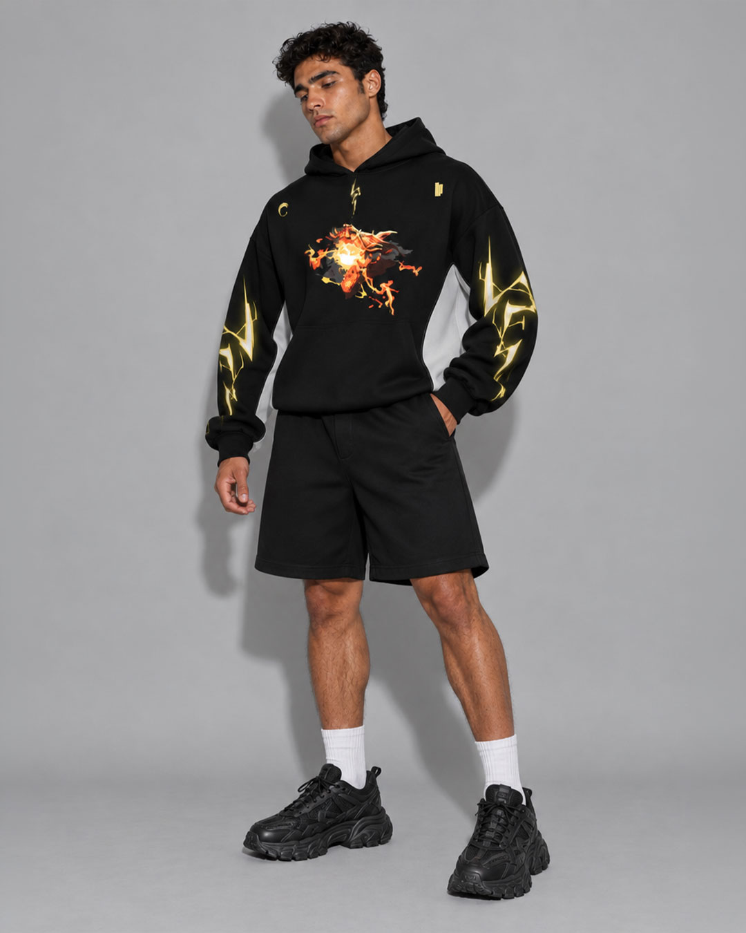 Model wearing a black hoodie with a fiery flame graphic on the chest and yellow lightning bolt print down the sleeves, paired with black shorts and black chunky sneakers — streetwear brand hoodie collection.