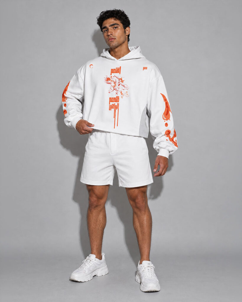 Male model wearing a white cropped hoodie with an orange dragon graphic and stacked script text on the chest, matching orange magma-splash sleeve prints, paired with matching white shorts, neutral gray studio background.