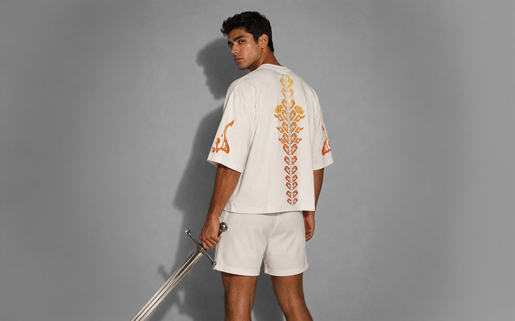 Back view of model wearing a white oversized t-shirt with an ombré orange-to-gold geometric spine print down the back and Arabic calligraphy sleeve prints, paired with white shorts, holding a sword — streetwear brand t-shirt collection.