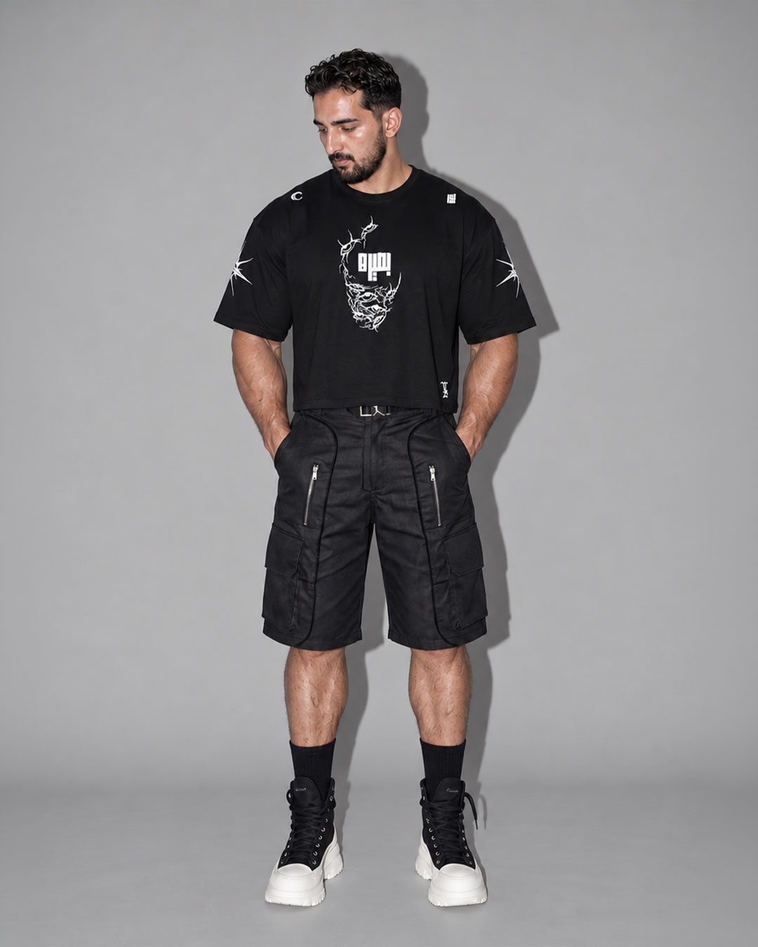 Male model wearing a black cropped t-shirt with a white skull-face graphic and stylized text on the chest, paired with black cargo shorts, black knee-high socks, and black-and-white platform sneakers, neutral gray studio background.