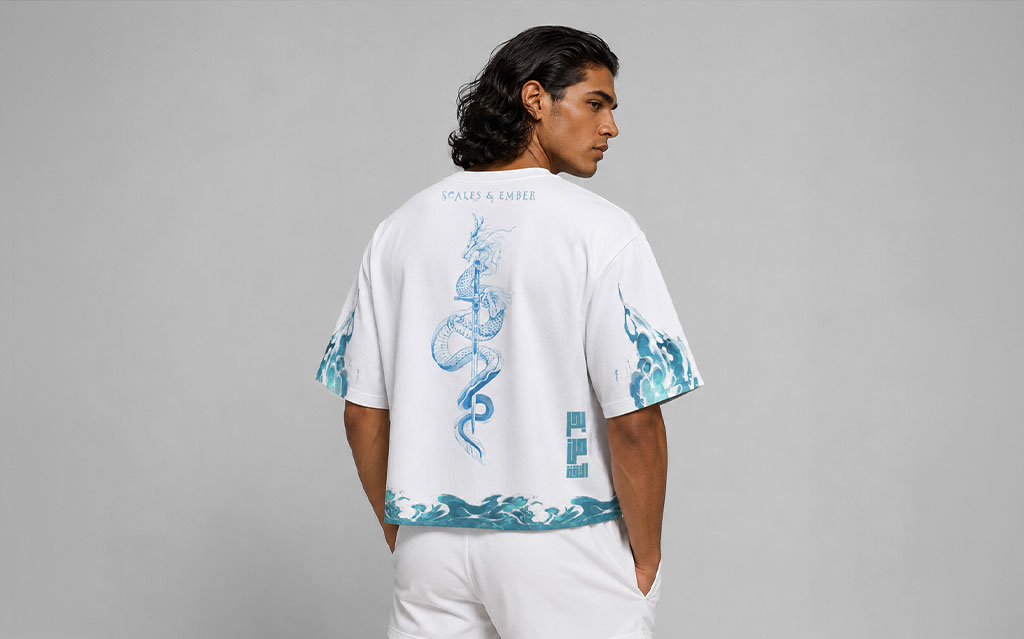 Back view of model wearing a white oversized t-shirt with a turquoise serpentine dragon graphic down the spine, "Scales & Ember" branding, wave hem print, and matching sleeve prints, paired with white shorts — streetwear brand t-shirt collection.