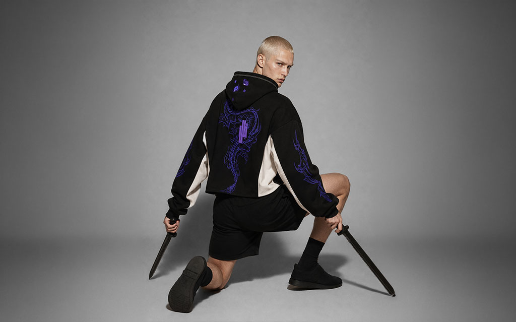 Back view of model wearing a black cropped hoodie with a purple serpentine dragon graphic down the back, flame sleeve prints, cream side panels, paired with black shorts and black boots, holding a sword — streetwear brand hoodie collection.