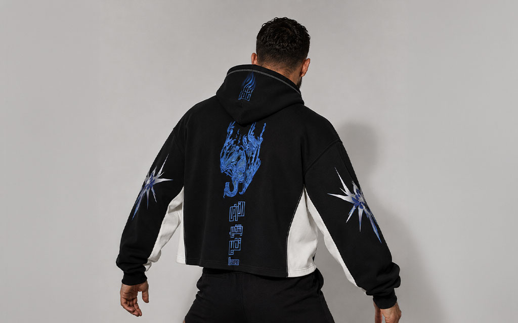 Back view of model wearing a black cropped hoodie with a blue flame-dragon graphic and Arabic calligraphy print on the back, starburst sleeve prints, white side panels — streetwear brand hoodie collection.