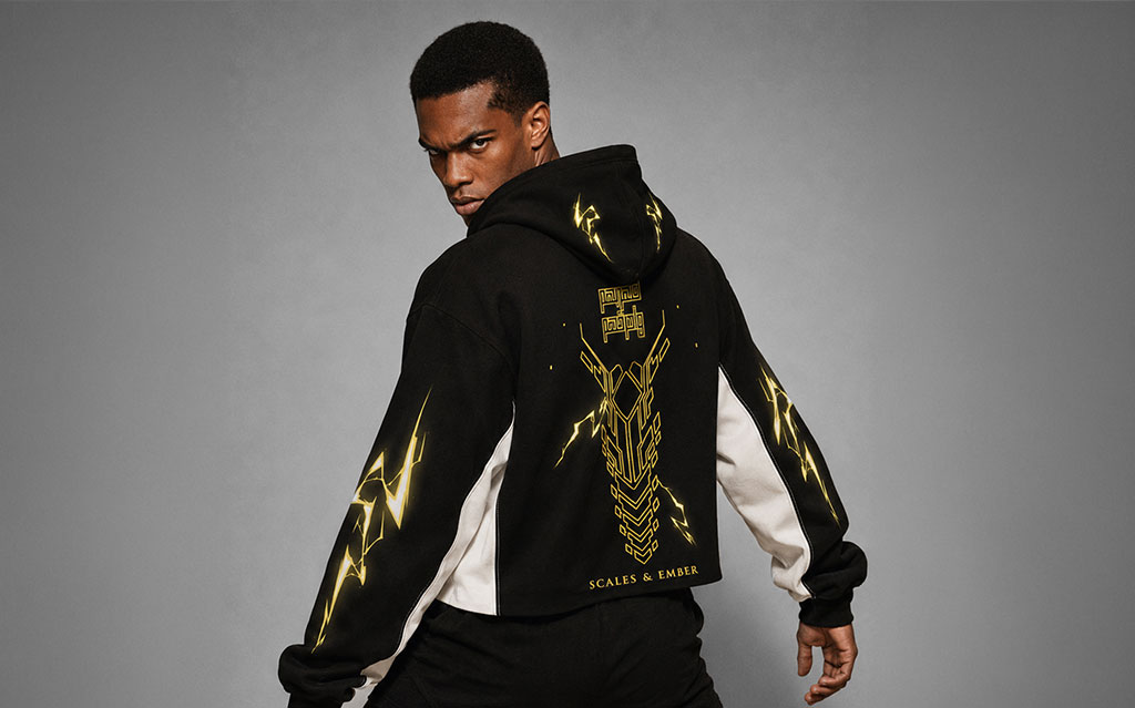 Back view of model wearing a black cropped hoodie with a gold lightning dragon-head graphic and "Scales & Ember" branding on the back, lightning bolt sleeve prints, white side panels, paired with black shorts — streetwear brand hoodie collection.